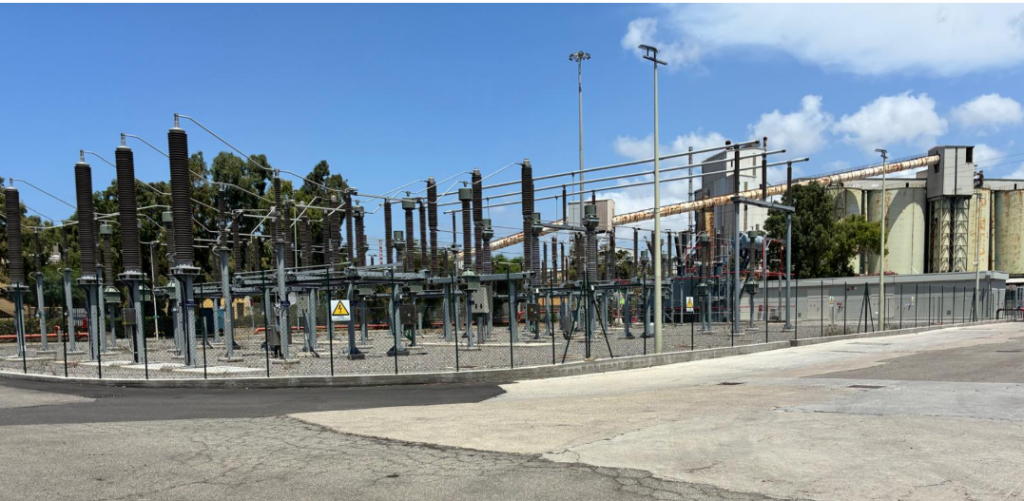 230/33 kV AIS electrical substation serving the BESS plants at the Portoscuso and Sulcis power stations, with insulators, gantries and conductors behind the perimeter fence and the industrial plant in the background.