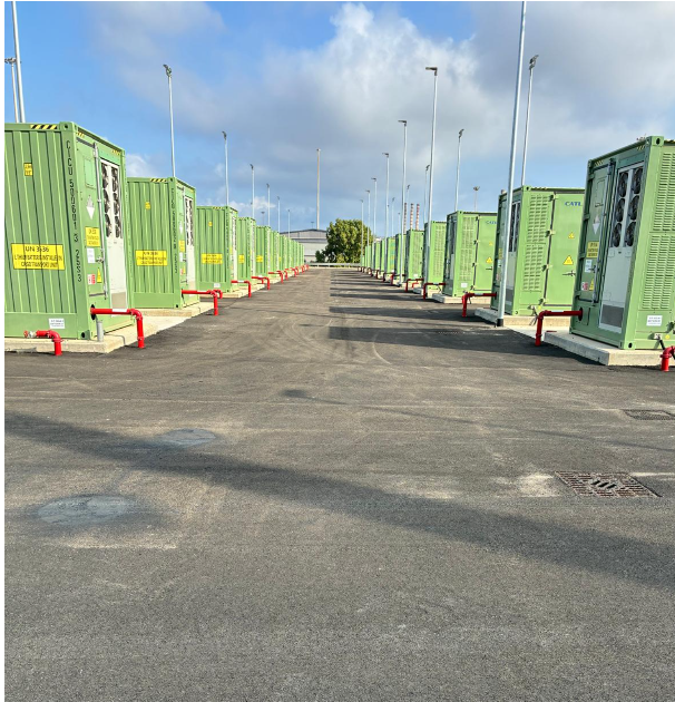 Row of battery containers at the BESS plant of the Enel Sulcis power station in Portoscuso, aligned along the paved access road with the power station's chimneys in the background.